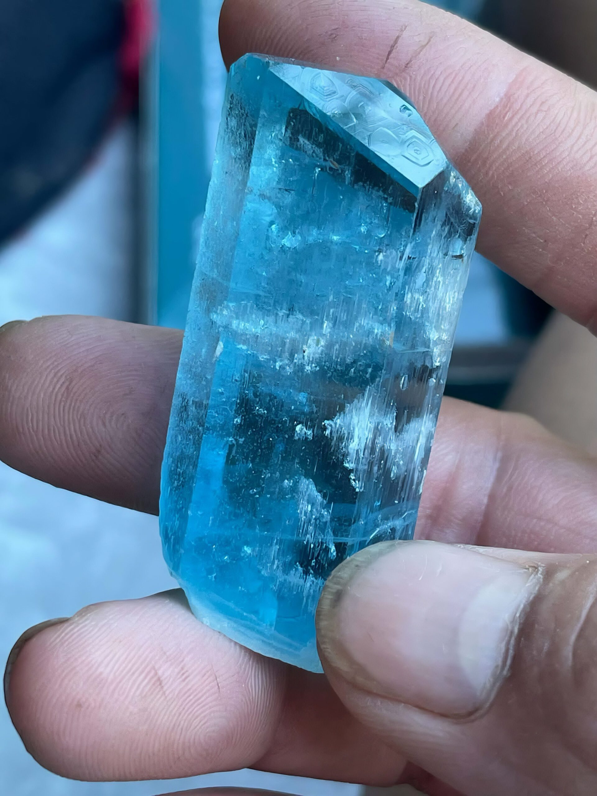 Blue aquamarine crystal held between fingers, showing internal detail and geometric markings on its termination
